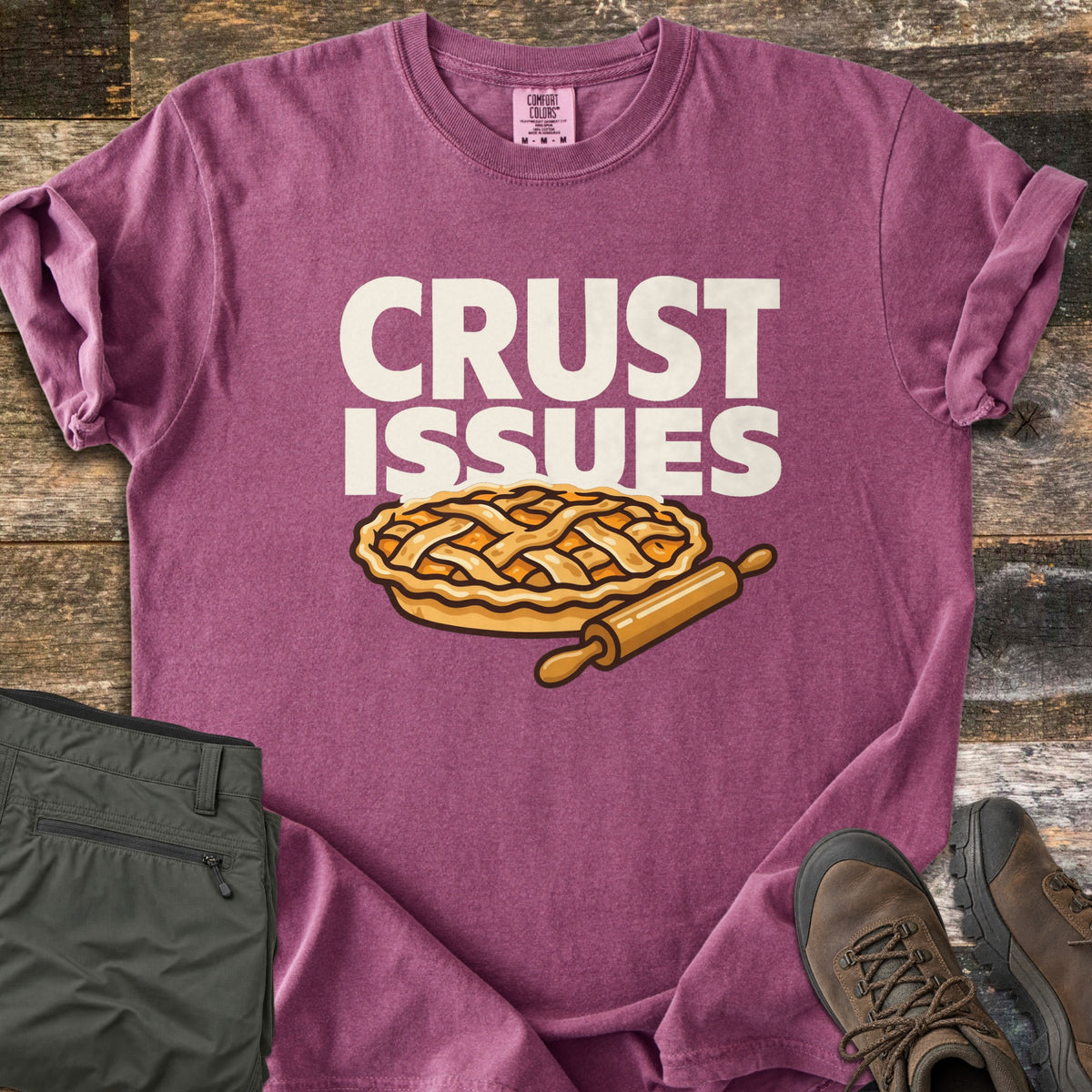 Crust Issues Comfort Colors T-shirt in Berry, laid flat. The words CRUST ISSUES in off-white block letters sit above a cartoon lattice-top pie and a wooden rolling pin. Olive pants and brown hiking boots lie beside it on a rustic wood surface.