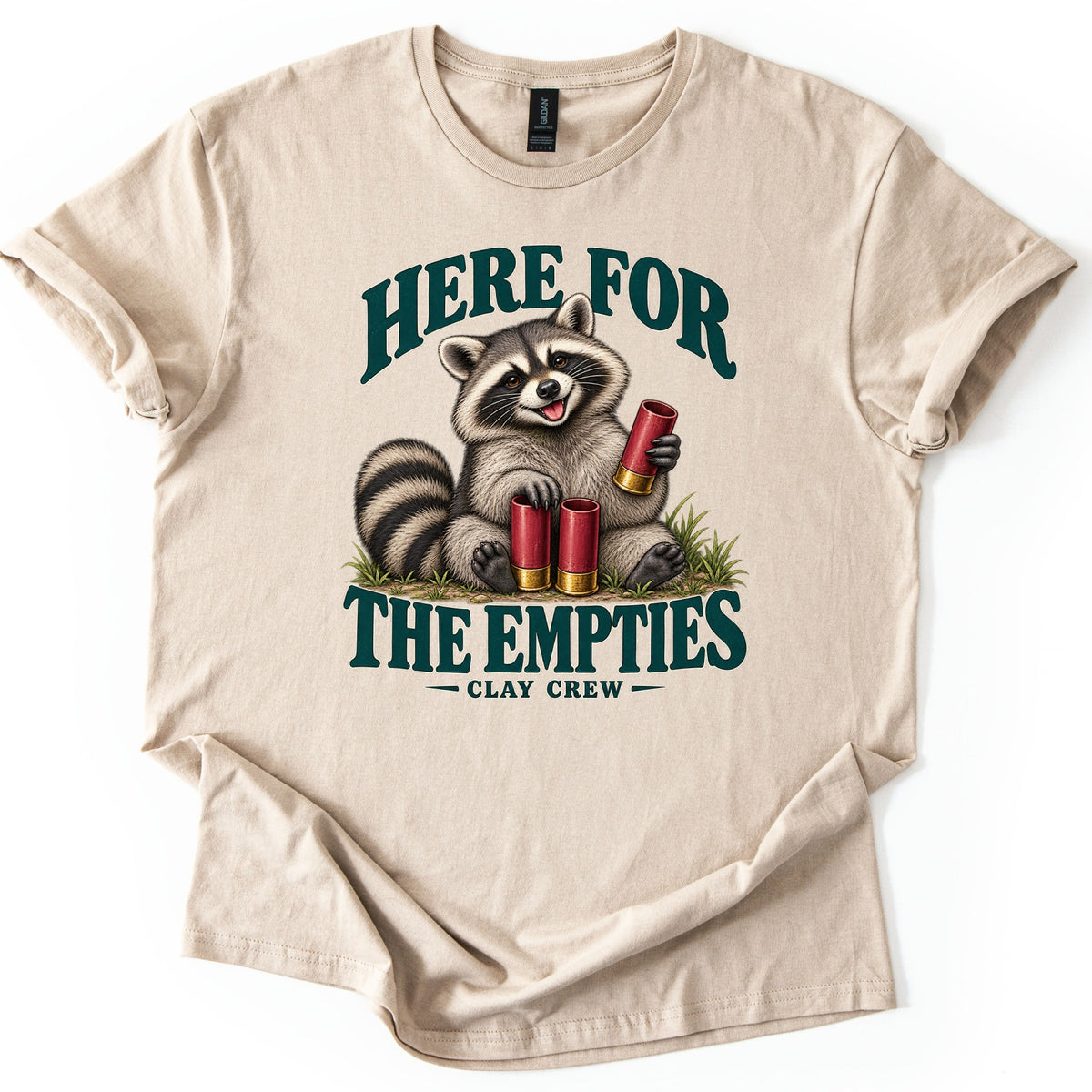 Here for the Empties Comfort Colors T-shirt in Sand, laid flat. A grinning raccoon sits in grass holding a red shotgun shell, with two more at its feet, between the words Here For The Empties and Clay Crew in dark green. Sleeves are cuffed and the hem folded, on white.