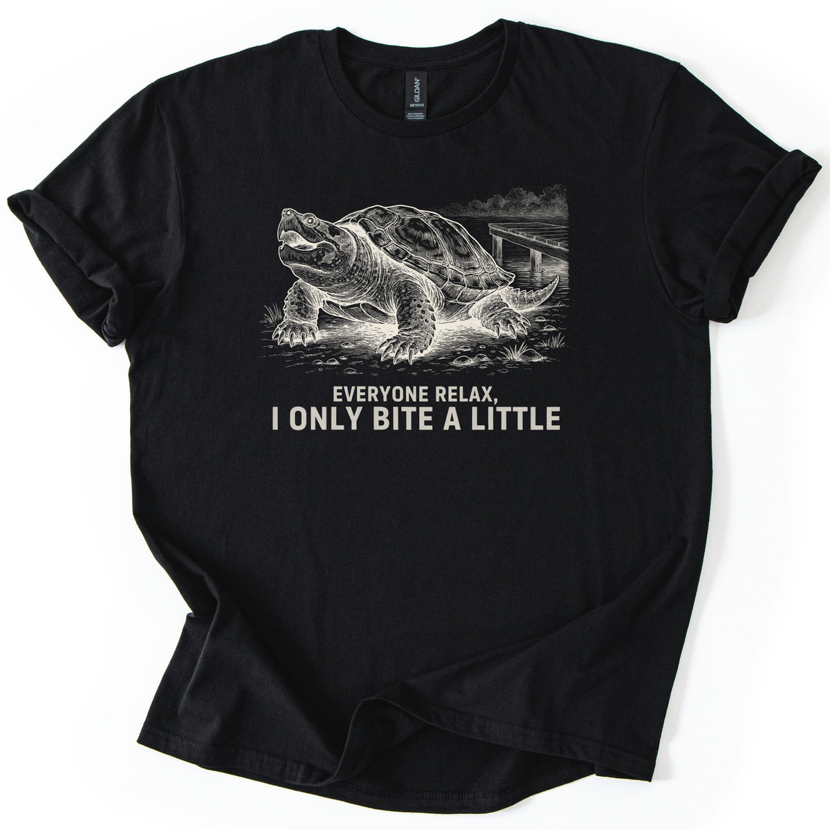 Everyone Relax I Only Bite A Little Comfort Colors T-shirt in Black, laid flat. A cream ink drawing of a snapping turtle with its mouth open on a shoreline by a wooden dock, above the words Everyone Relax, I Only Bite A Little. Sleeves are rolled on a white background.