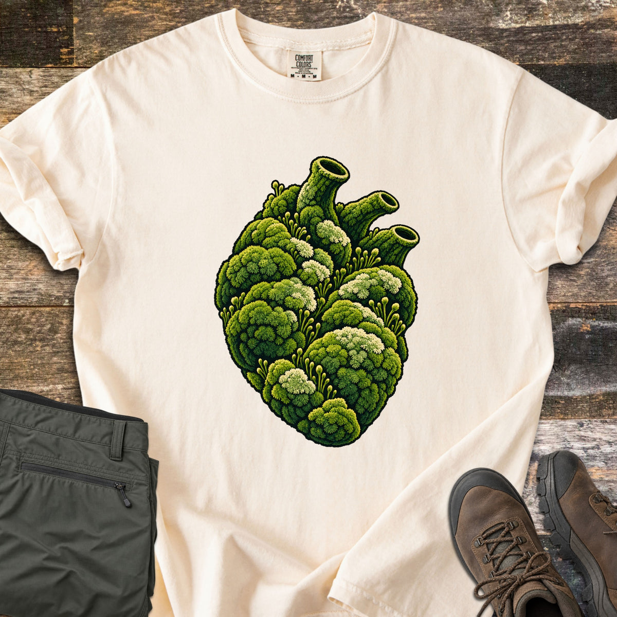 Moss Heart Comfort Colors T-shirt in Ivory, laid flat. An anatomical heart covered in green moss with pale tufts, its vessels shaped like moss-covered tubes. The shirt has rolled sleeves and lies on weathered wood beside folded olive pants and brown hiking boots.