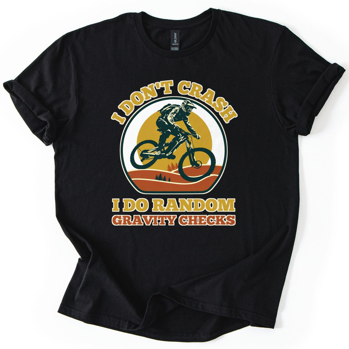 Random Gravity Checks MTB Comfort Colors T-shirt in Black, laid flat. A mountain biker in a full-face helmet jumps against a mustard sun over orange hills with pine trees, framed by the words I Don't Crash above and I Do Random Gravity Checks below. Sleeves rolled, on white.