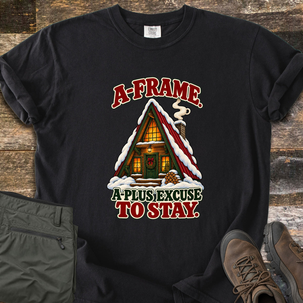 A-Frame A-Plus Excuse to Stay Comfort Colors T-shirt in Black, laid flat. A snowy red-roofed A-frame cabin with glowing windows, a wreathed green door, smoking chimney and stacked firewood, between the words A-Frame. A-Plus Excuse to Stay. Olive pants and brown boots lie on weathered wood.