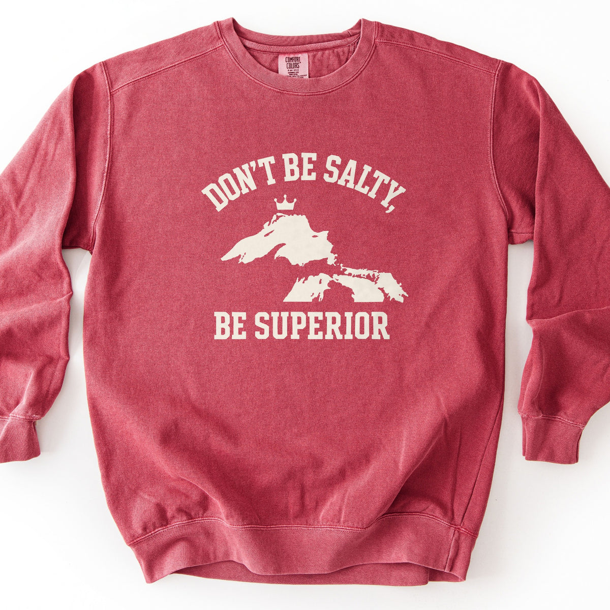 Be Superior Comfort Colors crewneck sweatshirt in Crimson, laid flat. Cream print of the words Don't Be Salty, arched above a silhouette of Lake Superior topped with a small crown, with Be Superior below. Laid on a white background.
