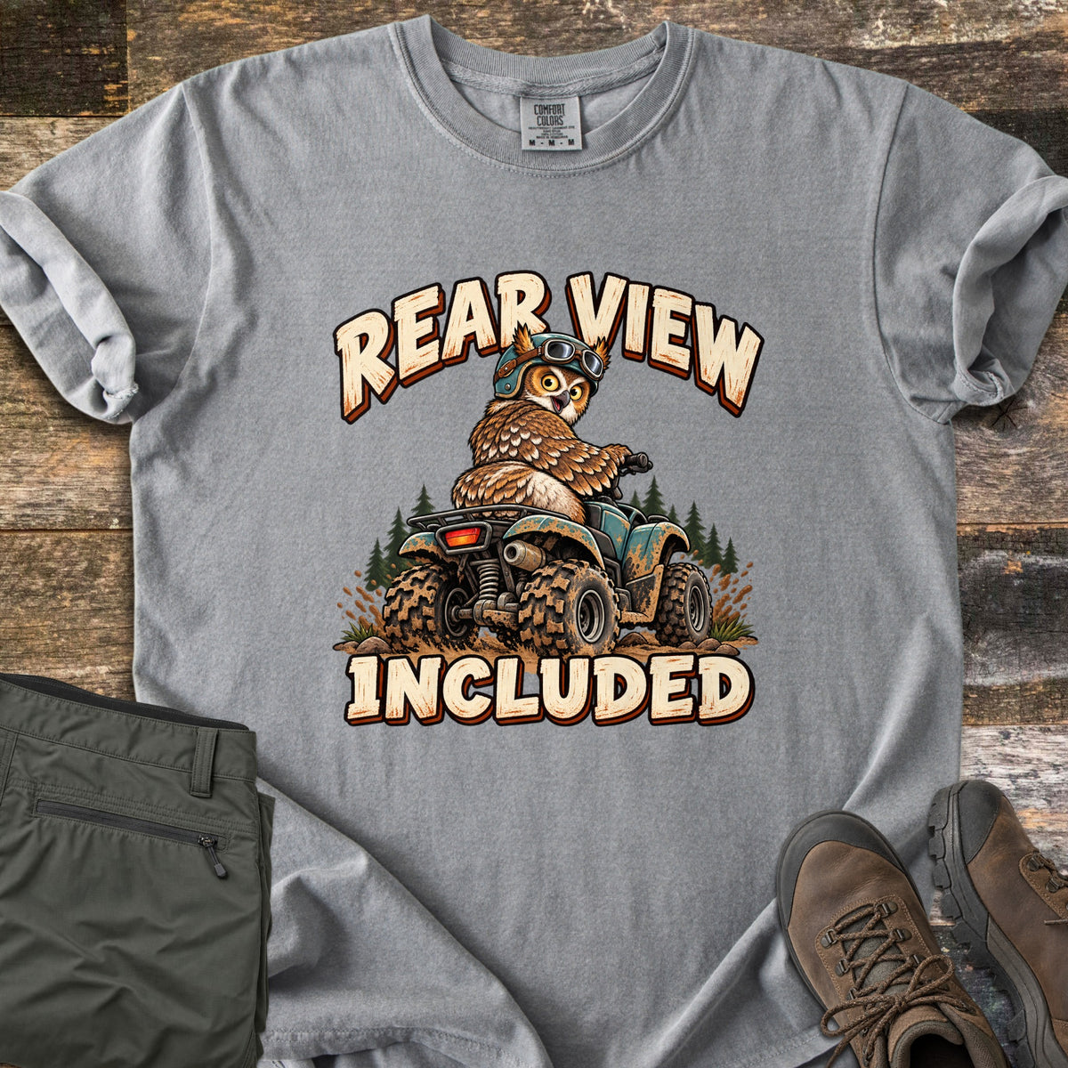 Rear View Included Comfort Colors T-shirt in Grey, laid flat. A cartoon owl in a helmet and goggles rides a muddy ATV past pine trees, looking back over its shoulder, between the words Rear View Included. Olive cargo pants and brown hiking boots lie on a wood floor.