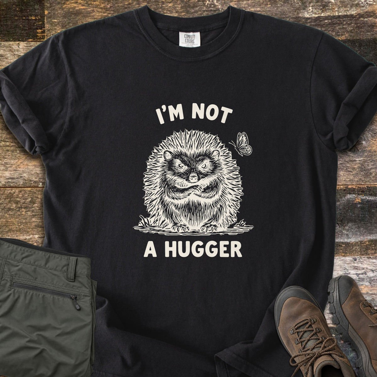 I'm Not A Hugger Comfort Colors T-shirt in Black, laid flat. A sketched grumpy hedgehog with crossed arms and a butterfly beside it, between the words I'M NOT and A HUGGER in cream. It lies on weathered wood with folded olive pants and brown hiking boots.