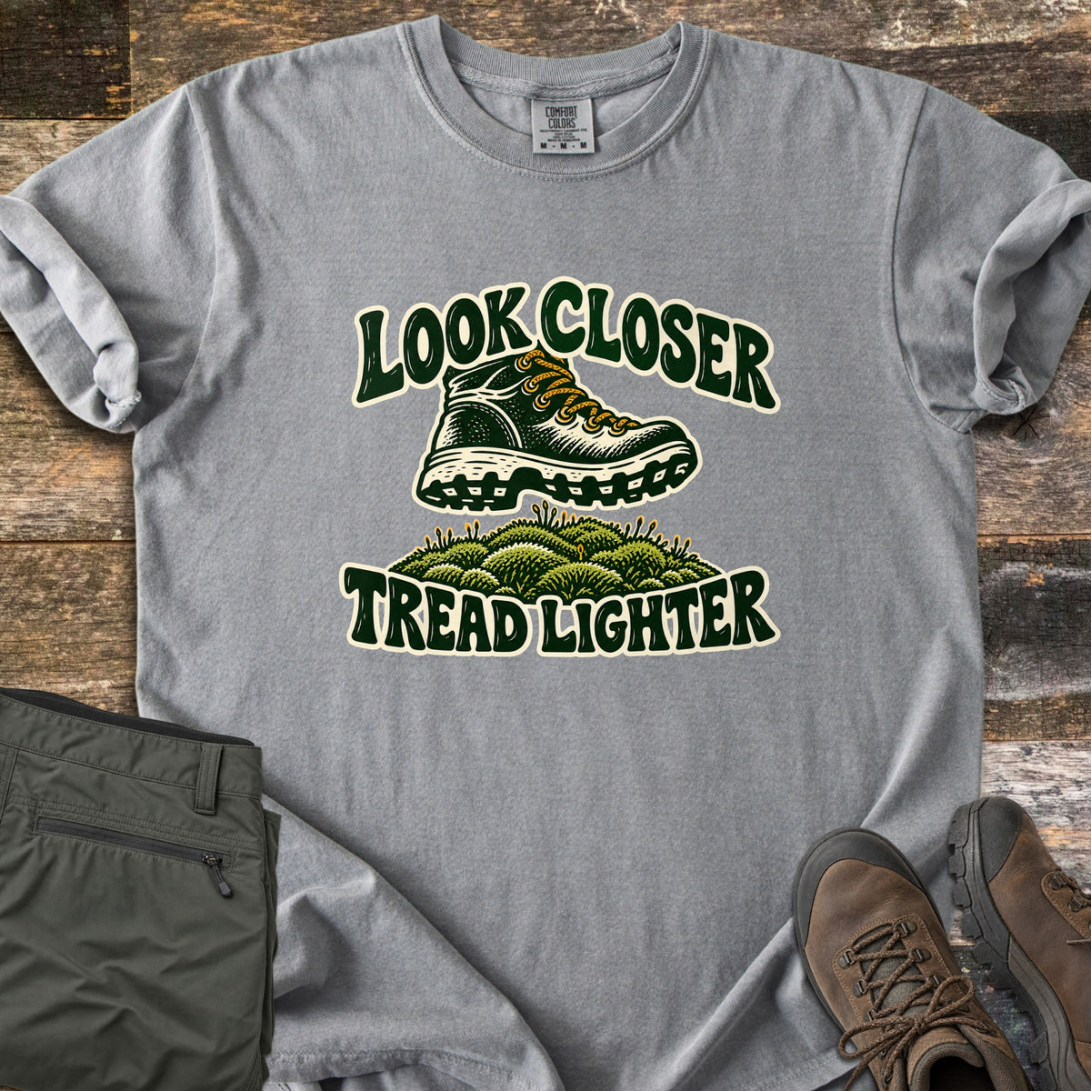 Look Closer Tread Lighter Comfort Colors T-shirt in Grey, laid flat. A green hiking boot with yellow laces steps above a mound of moss between the words Look Closer and Tread Lighter. Sleeves rolled, on weathered wood with folded olive hiking pants and brown boots.