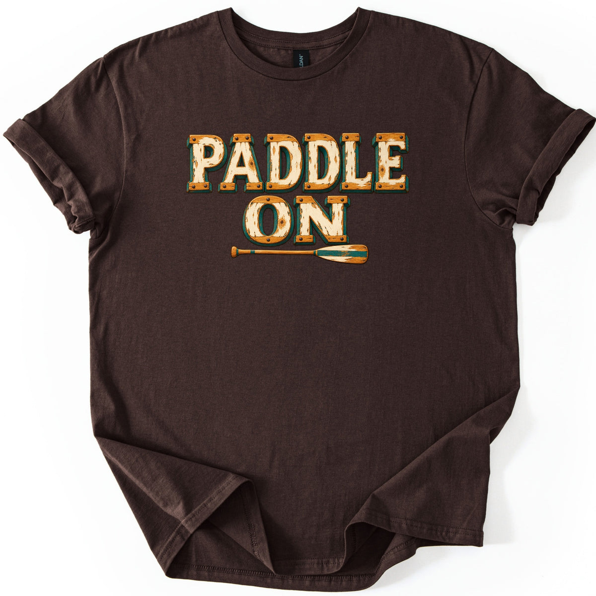 Paddle On Comfort Colors T-shirt in Dark Chocolate, laid flat. Weathered cream and orange wood-plank letters with rivets and green shadows spell PADDLE ON over a wooden canoe paddle with green stripes. The shirt lies with cuffed sleeves on a white background.