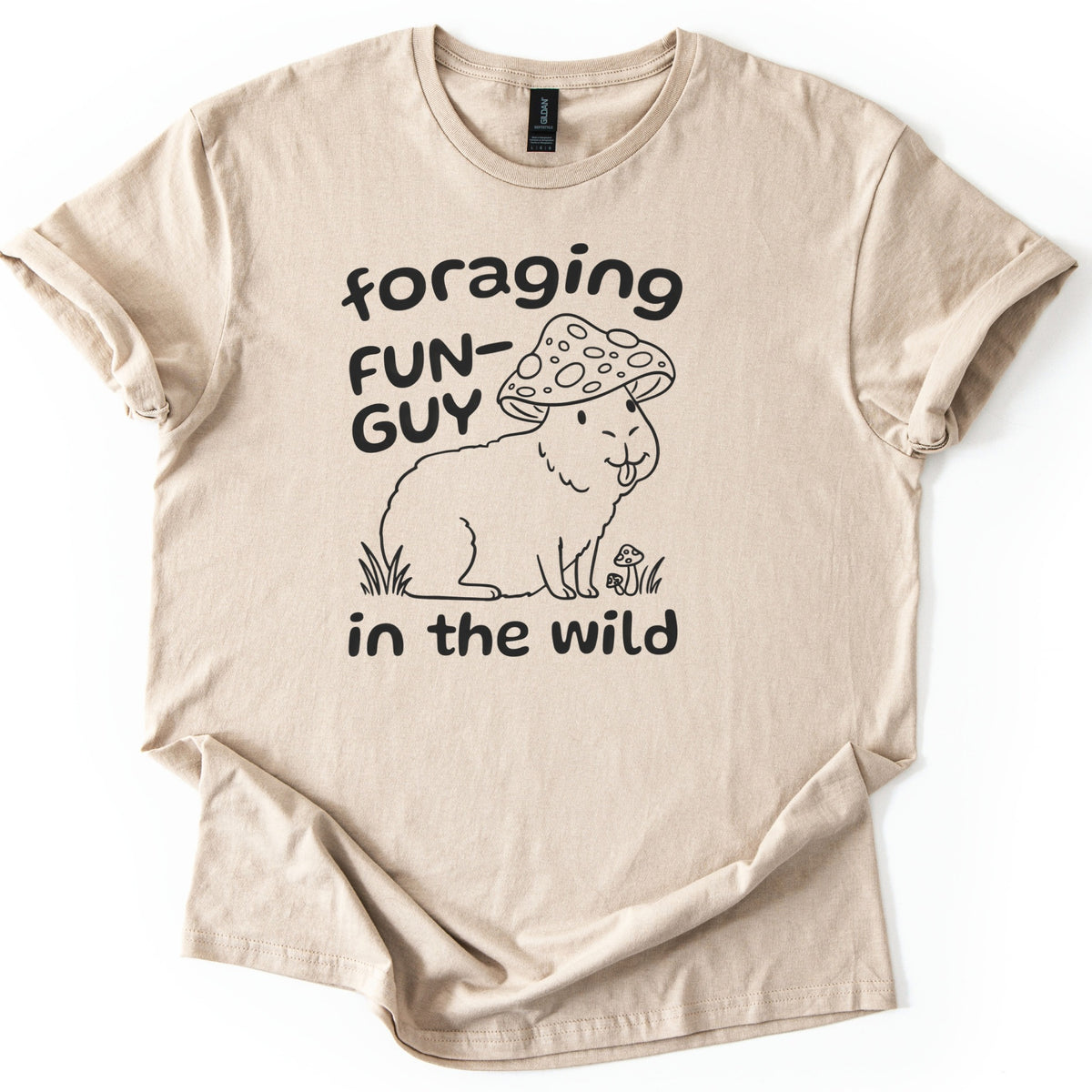 Foraging Fun-Guy Capybara Comfort Colors T-shirt in Sand, laid flat. A black line drawing of a winking capybara with its tongue out, wearing a spotted mushroom cap and sitting in grass by tiny mushrooms, with the words foraging FUN-GUY in the wild. Sleeves rolled, on white.