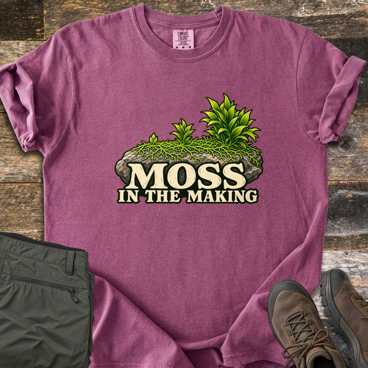 Moss in the Making Comfort Colors T-shirt in Berry, laid flat. A mossy gray rock sprouting green leafy plants sits above the words Moss in the Making. Olive hiking pants and brown hiking boots lie beside it on weathered wood planks.