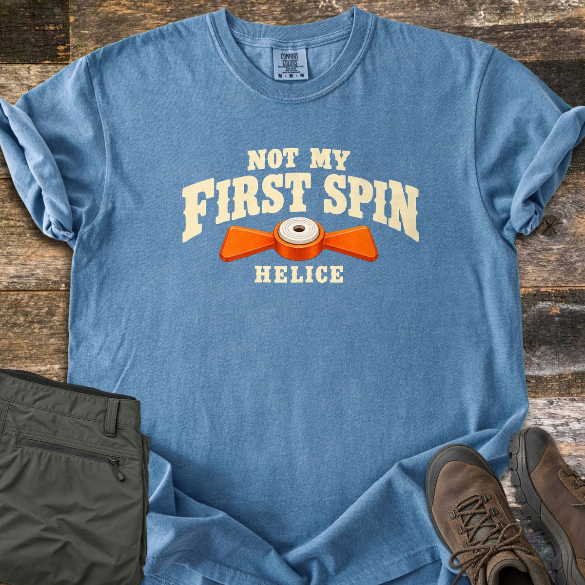 Not My First Spin Comfort Colors T-shirt in Blue Jean, laid flat. Cream arched text reads Not My First Spin above an orange two-winged helice target with a white center, with Helice below. Folded olive pants and brown hiking boots sit beside it on weathered wood planks.
