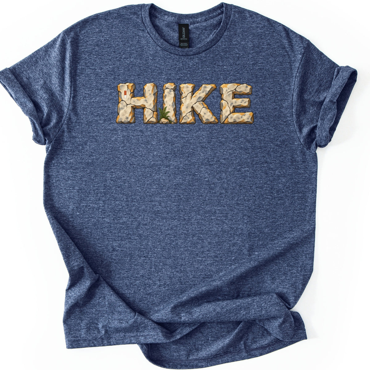 Hike Comfort Colors T-shirt in Heather Navy, laid flat. The word HIKE is printed in letters made of cracked tan stone with orange edges, a small green fern at the base and a red and white trail marker on the H. Sleeves rolled, on a white background.