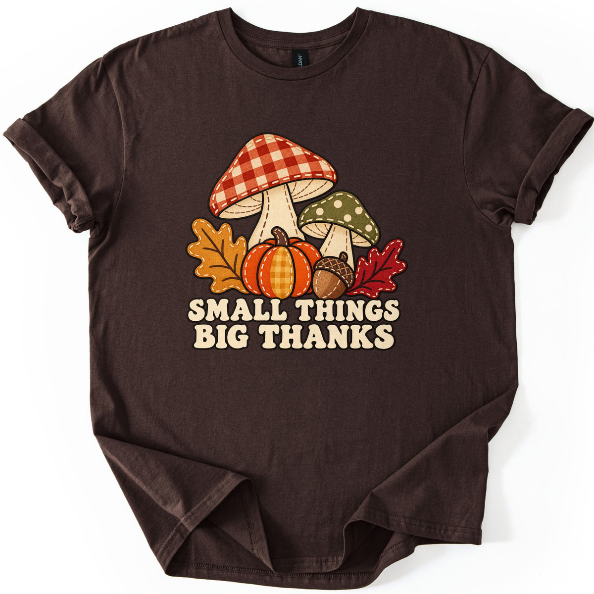Small Things Big Thanks Comfort Colors T-shirt in Dark Chocolate, laid flat. Stitched red gingham and green polka-dot mushrooms rise behind an orange pumpkin, an acorn and orange and red leaves above the words Small Things Big Thanks. Sleeves rolled, on a white backdrop.