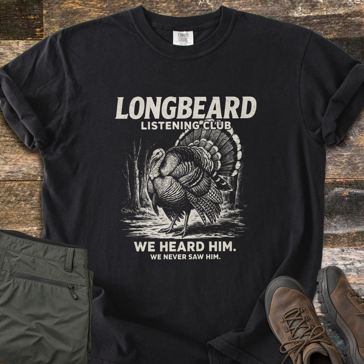 Longbeard Listening Club Comfort Colors T-shirt in Black, laid flat. Cream text reads Longbeard Listening Club over an engraved wild turkey strutting in the woods, then We heard him. We never saw him. Folded olive hiking pants and brown boots rest beside it on weathered wood.