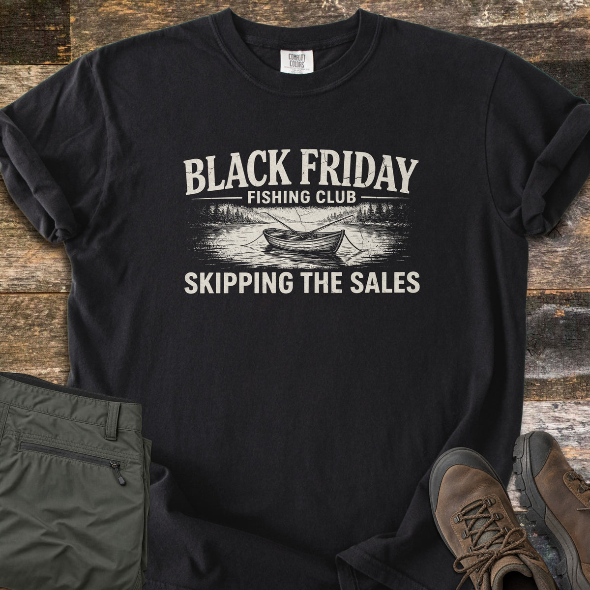 Black Friday Fishing Club Comfort Colors T-shirt in Black, laid flat. A distressed cream print shows the words Black Friday Fishing Club above a rowboat with oars on a lake lined with pine trees, and Skipping the Sales below. Olive pants and brown hiking boots sit beside it on wood planks.