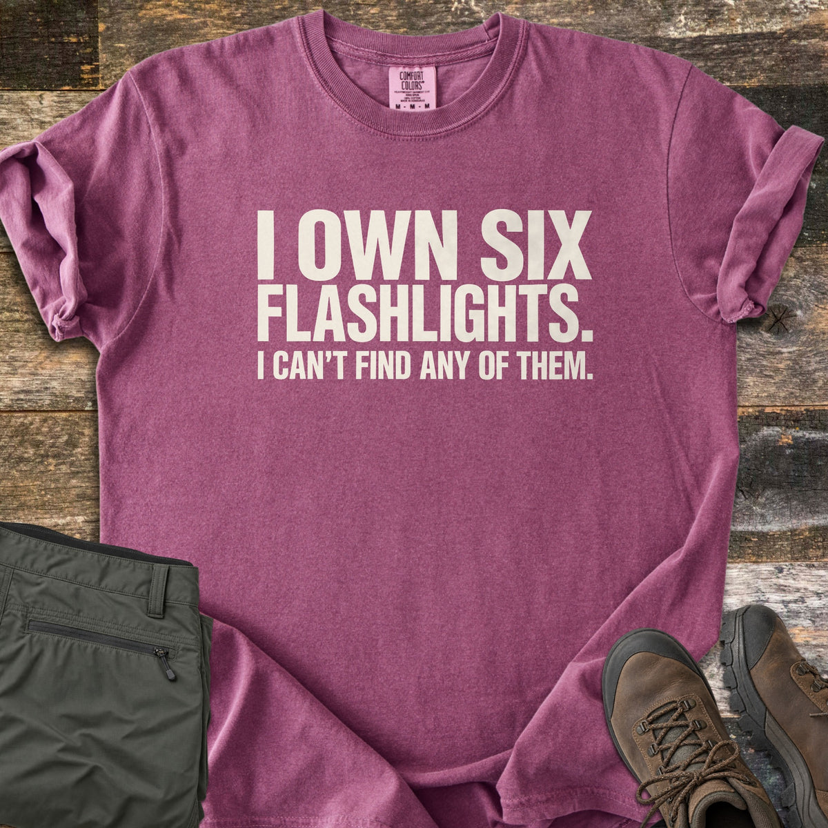 Six Flashlights Comfort Colors T-shirt in Berry, laid flat. Bold off-white text reads I OWN SIX FLASHLIGHTS. I CAN'T FIND ANY OF THEM. The shirt has rolled sleeves and lies on weathered wood beside folded olive cargo pants and brown hiking boots.