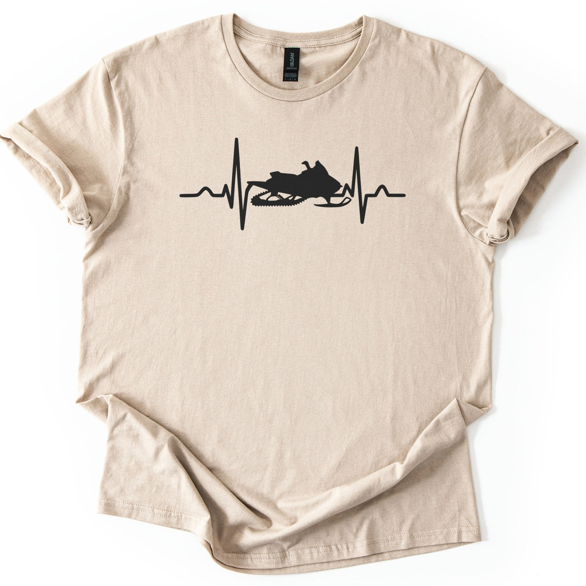 Heartbeat Snowmobile Comfort Colors T-shirt in Sand, laid flat. A black heartbeat line spans the chest with a snowmobile silhouette in the middle of the pulse, with no words. The shirt has rolled sleeves and lies on a white background.