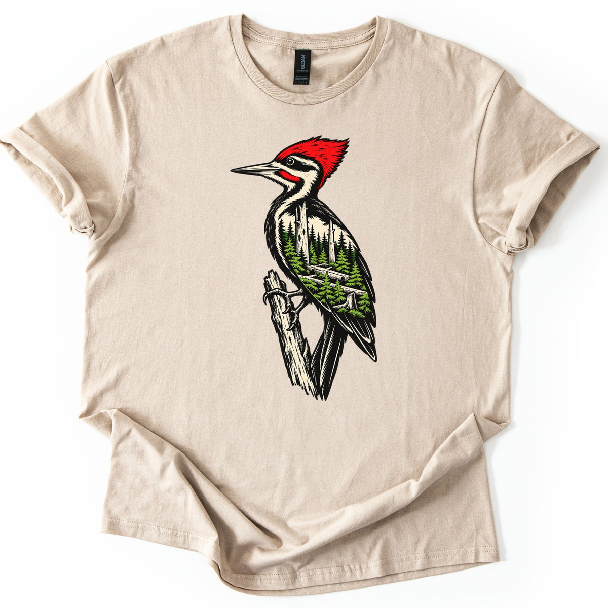 Woodpecker Forest Comfort Colors T-shirt in Sand, laid flat. A woodpecker with a red crest sits on a broken branch, its black and cream feathers holding a scene of evergreen trees. No words are printed. The sleeves are cuffed and the shirt lies on a white background.