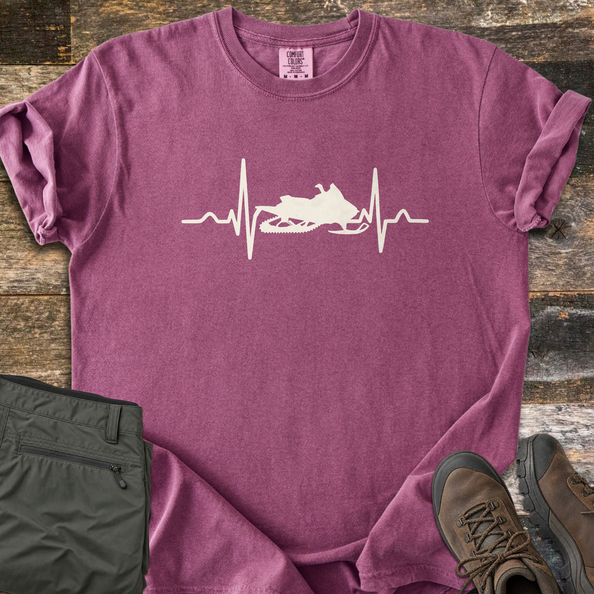 Snowmobile Heartbeat Comfort Colors T-shirt in Berry, laid flat. A cream heartbeat line runs across the chest with a snowmobile silhouette in its center, with no words. Sleeves are rolled, on weathered wood planks beside folded olive pants and brown hiking boots.