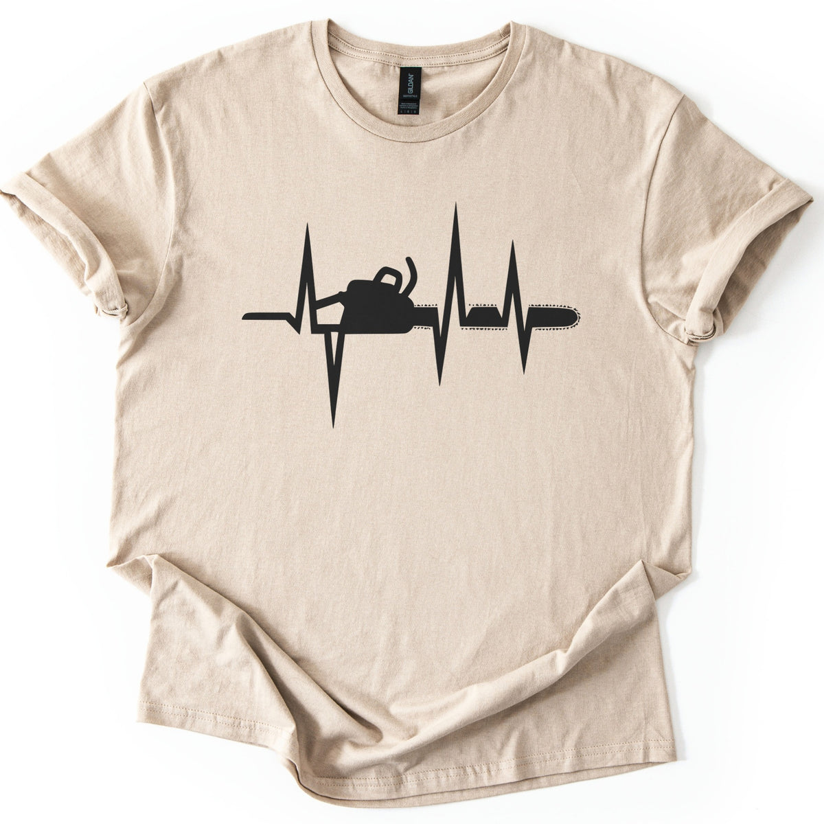 Chainsaw Heartbeat Comfort Colors T-shirt in Sand, laid flat. A black heartbeat line with sharp peaks runs across the chest, with a chainsaw silhouette set into the line, its bar and chain extending to the right. The sleeves are rolled on a white background.