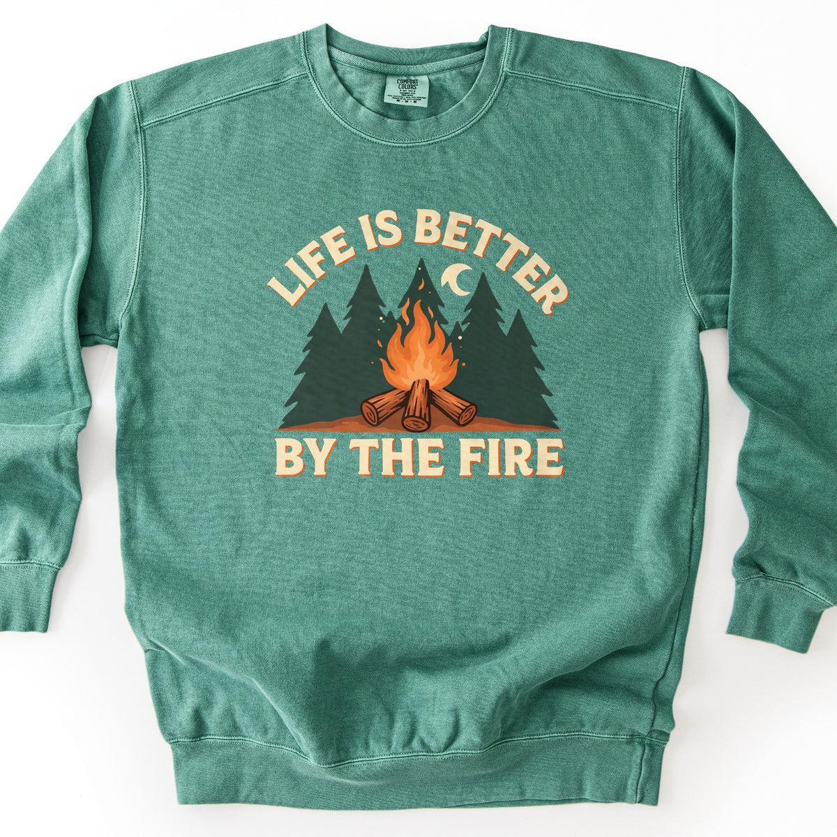 Life Is Better By The Fire Comfort Colors crewneck sweatshirt in Light Green, laid flat. Cream letters with an orange shadow arch Life Is Better over dark green pine trees, a crescent moon and a campfire of crossed logs, with By The Fire below. It lies on a white surface.