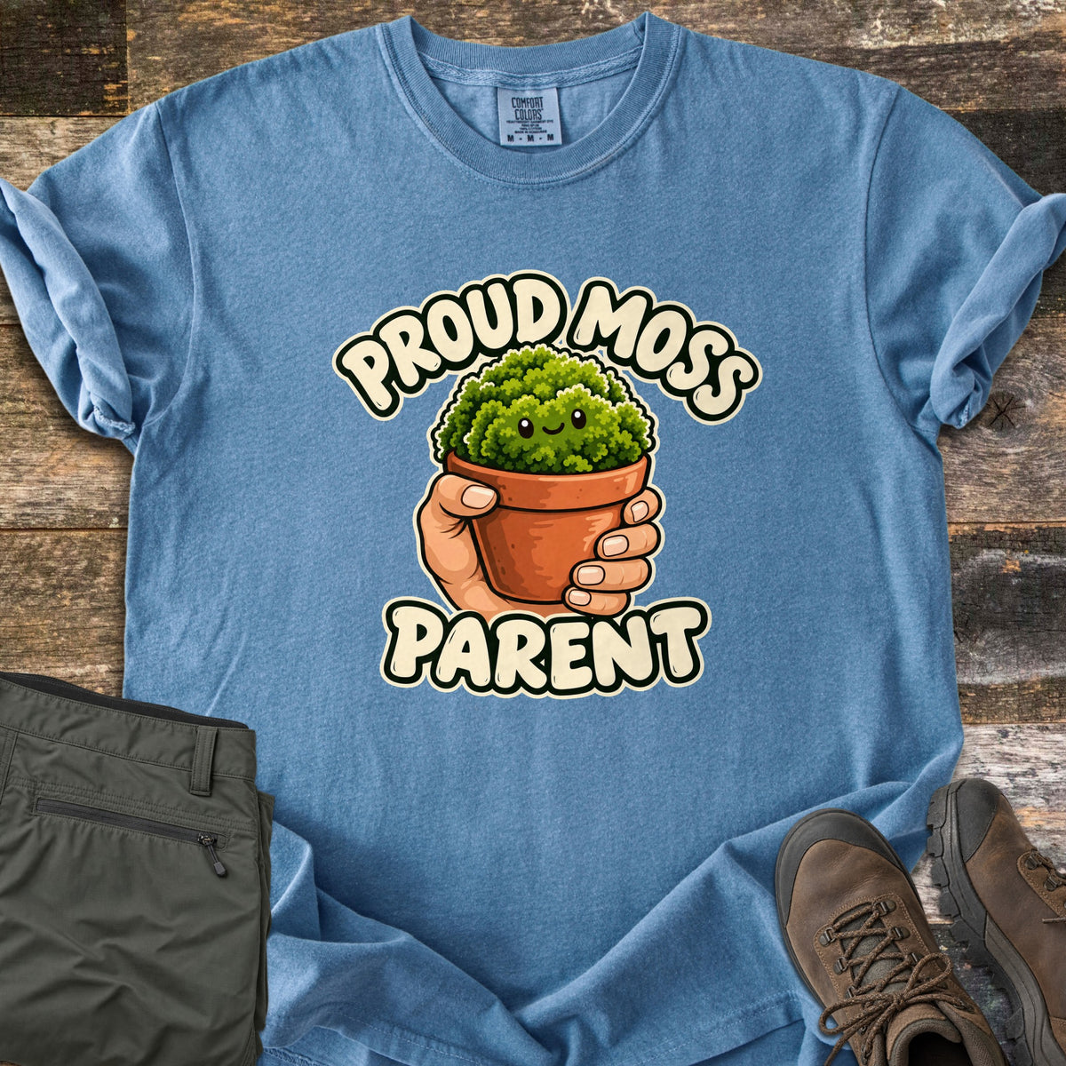 Proud Moss Parent Comfort Colors T-shirt in Blue Jean, laid flat. A cartoon hand holds a terracotta pot of smiling green moss between the words Proud Moss Parent. Sleeves are rolled, with green hiking pants and brown hiking boots on a weathered wood background.