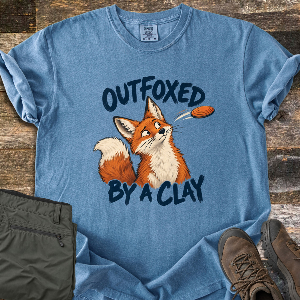 Outfoxed by a Clay Comfort Colors T-shirt in Blue Jean, laid flat. A cartoon red fox glances sideways at an orange clay target flying past, between the words Outfoxed By A Clay in navy brush letters. Sleeves rolled on rustic wood with olive pants and brown hiking boots.