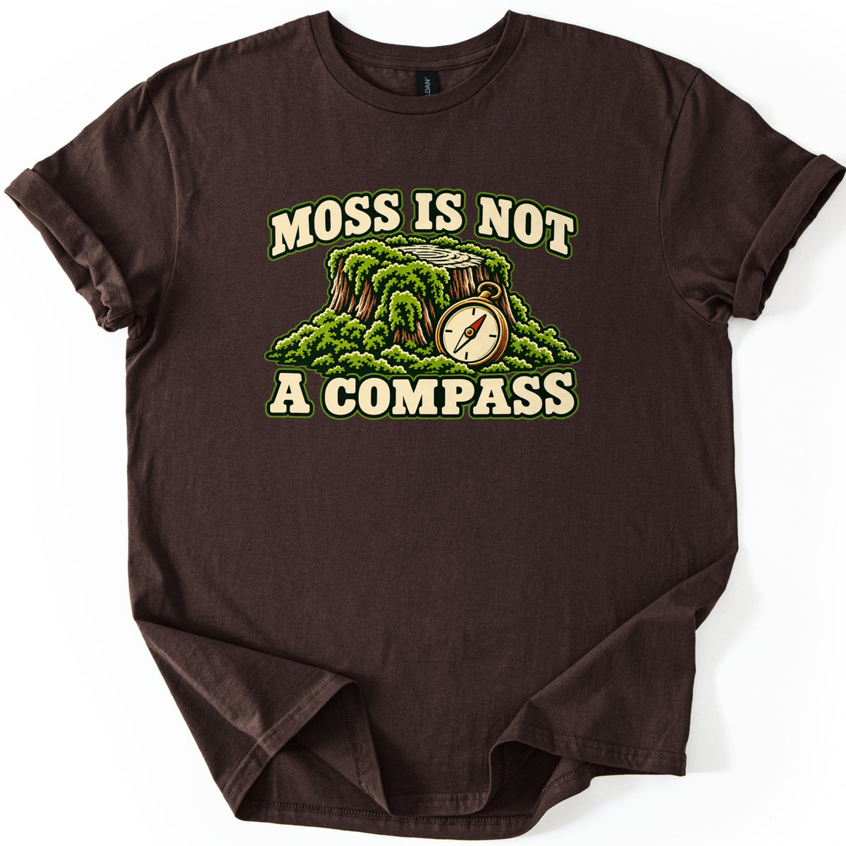 Moss Is Not a Compass Comfort Colors T-shirt in Dark Chocolate, laid flat. Moss Is Not is printed in arched cream letters over a moss-covered stump and a brass pocket compass, with A Compass underneath. The shirt lies flat on a white background with cuffed sleeves.