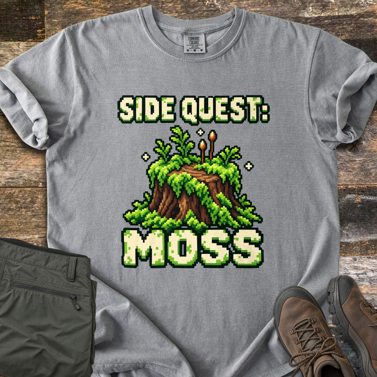 Side Quest Moss Comfort Colors T-shirt in Grey, laid flat. Pixel art of a moss-covered tree stump with ferns, two small sporophytes and sparkles, between the words Side Quest: and Moss. Olive cargo pants and brown hiking boots lie beside it on weathered wood planks.