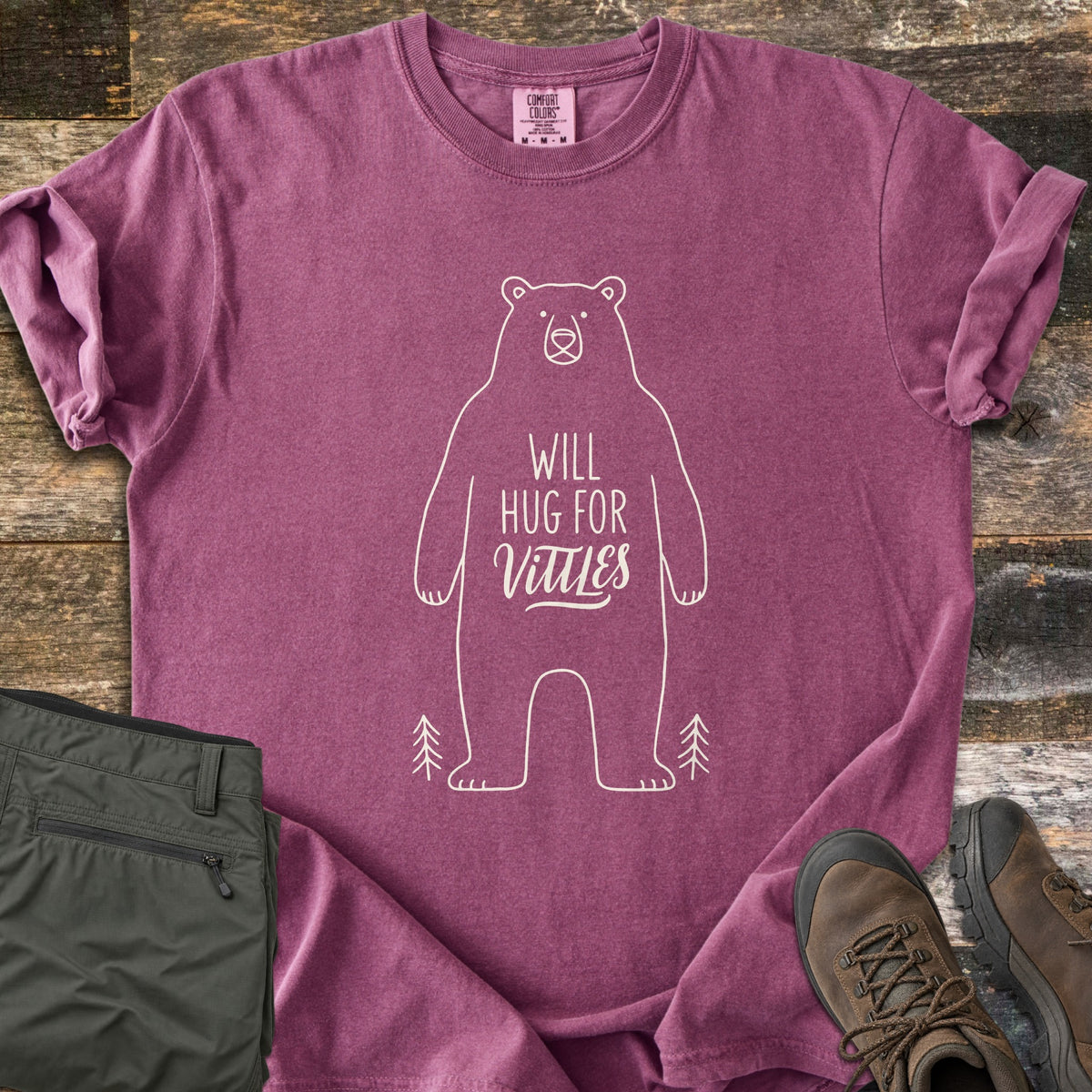 Will Hug For Vittles Comfort Colors T-shirt in Berry, laid flat. A cream outline of a standing bear has the words Will Hug For Vittles on its belly, with two small pine trees by its feet. Folded olive hiking pants and brown hiking boots sit beside it on weathered wood.