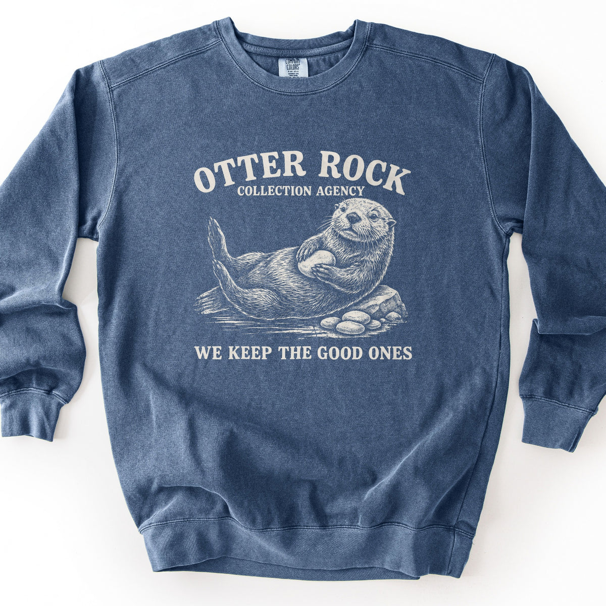 Otter Rock Collection Agency Comfort Colors crewneck sweatshirt in Denim, laid flat. A cream sketch-style sea otter floats on its back hugging a smooth stone beside a rock and pebbles, under the words Otter Rock Collection Agency and above We Keep The Good Ones. Shown on a white background.