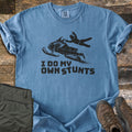 I Do My Own Stunts Comfort Colors T-shirt in Blue Jean, laid flat. A black silhouette of a rider flying off a jumping snowmobile, snow spraying behind, above the words I Do My Own Stunts. Folded olive cargo pants and brown hiking boots sit beside it on weathered wood.