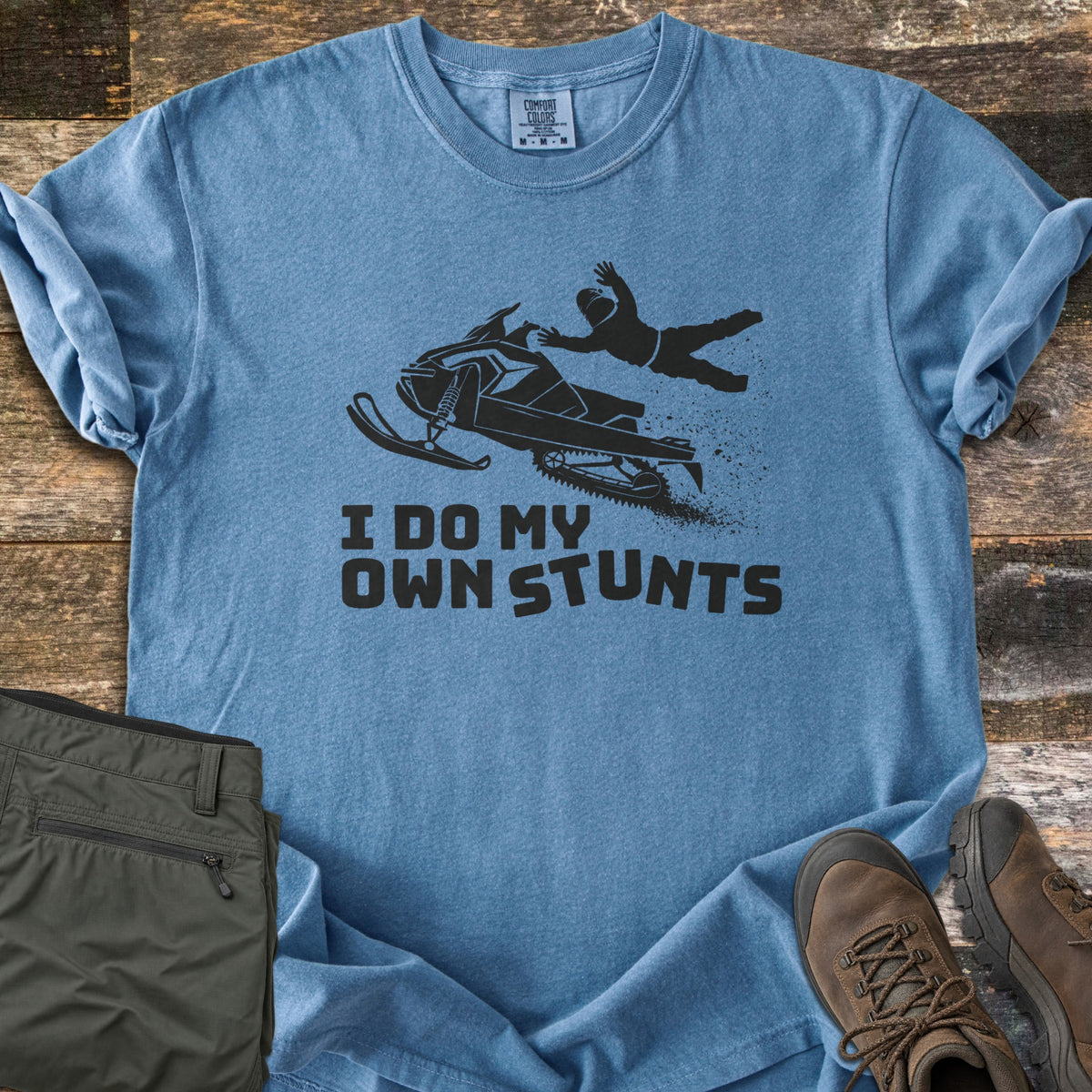 I Do My Own Stunts Comfort Colors T-shirt in Blue Jean, laid flat. A black silhouette of a rider flying off a jumping snowmobile, snow spraying behind, above the words I Do My Own Stunts. Folded olive cargo pants and brown hiking boots sit beside it on weathered wood.
