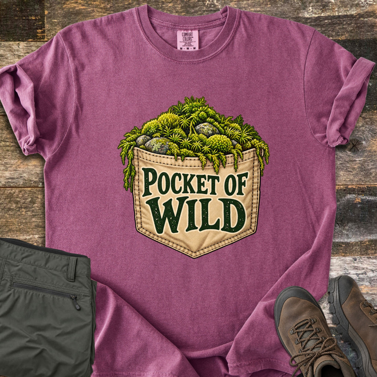 Pocket of Wild Comfort Colors T-shirt in Berry, laid flat. A tan stitched jeans pocket overflowing with green moss and lichen-covered stones, printed with the words Pocket of Wild. Shown on weathered wood with folded olive hiking pants and brown hiking boots.
