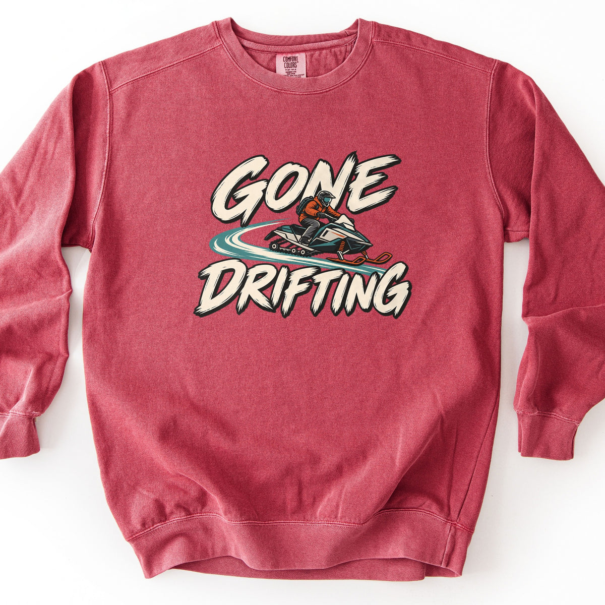 Gone Drifting Comfort Colors crewneck sweatshirt in Crimson, laid flat. A rider in an orange jacket and helmet drifts a teal and white snowmobile, kicking up a teal snow trail, between the words Gone Drifting in cream brush lettering. Shown on a white background.