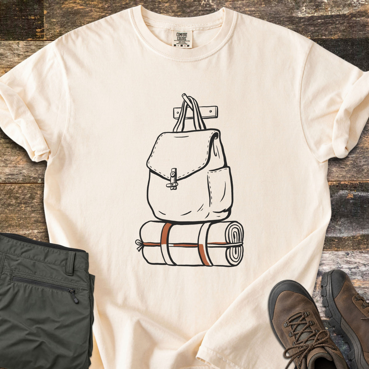 Backpack And Bedroll Comfort Colors T-shirt in Ivory, laid flat. Black line art of a canvas backpack hanging from a wall peg above a bedroll tied with rust-orange straps. Olive hiking pants and brown hiking boots sit beside it on weathered wood planks.