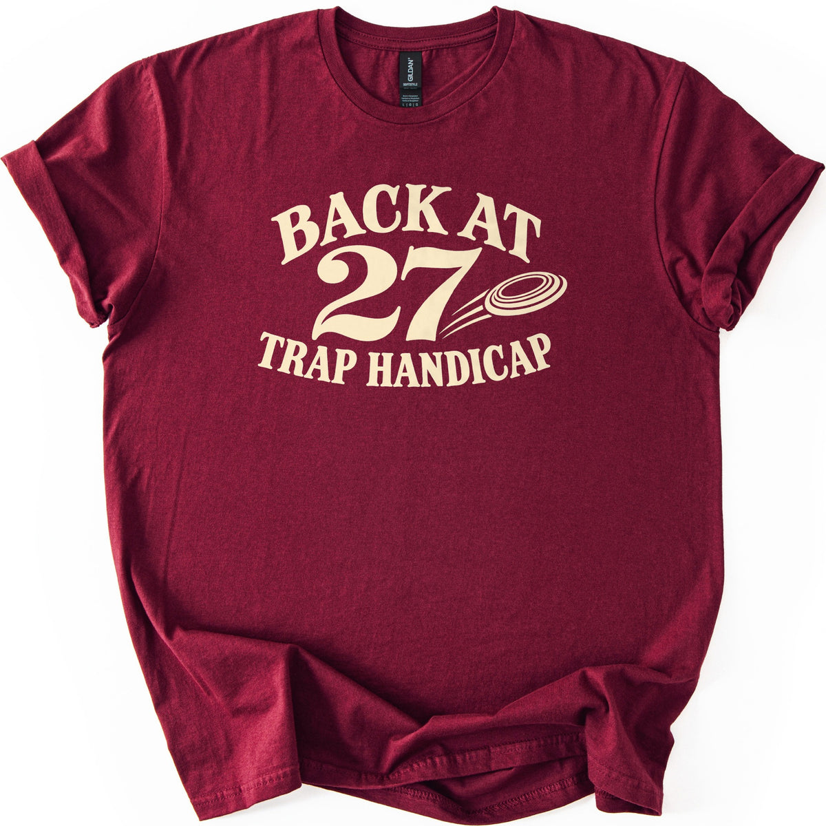 Back at 27 Trap Handicap Comfort Colors T-shirt in Maroon, laid flat. Cream lettering reads Back at 27 Trap Handicap, with a clay target trailing motion lines beside the large 27. The shirt has rolled sleeves and lies on a white background with a rumpled hem.