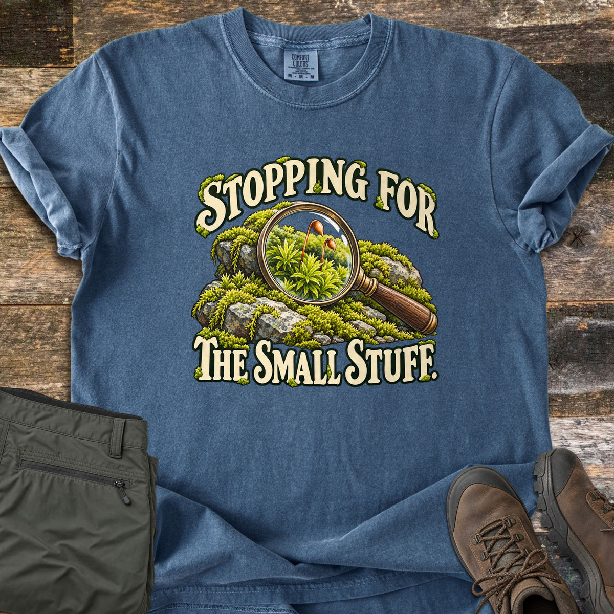Stopping for the Small Stuff Comfort Colors T-shirt in Denim, laid flat. A magnifying glass over a mossy rock reveals tiny moss sprouts, with the words Stopping for the Small Stuff. Shown on a wood floor with folded olive hiking pants and brown hiking boots.