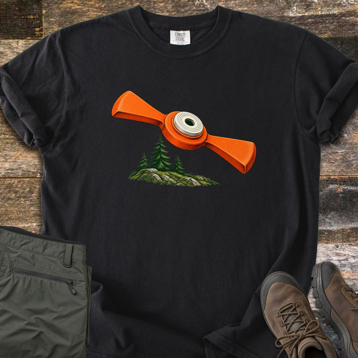Winged Target Over the Pines Comfort Colors T-shirt in Black, laid flat. An orange winged clay target with a white center flies over three pine trees on a mossy, rocky knoll, with no words. Folded olive hiking pants and brown hiking boots rest beside it on rustic wood planks.