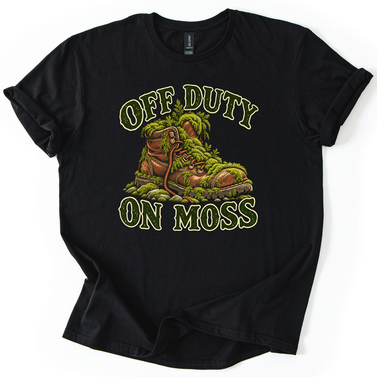 Off Duty on Moss Comfort Colors T-shirt in Black, laid flat. A worn brown leather hiking boot overgrown with green moss and ferns sits on mossy stones between the words OFF DUTY and ON MOSS in dark green letters with cream outlines. The shirt lies on a white surface.