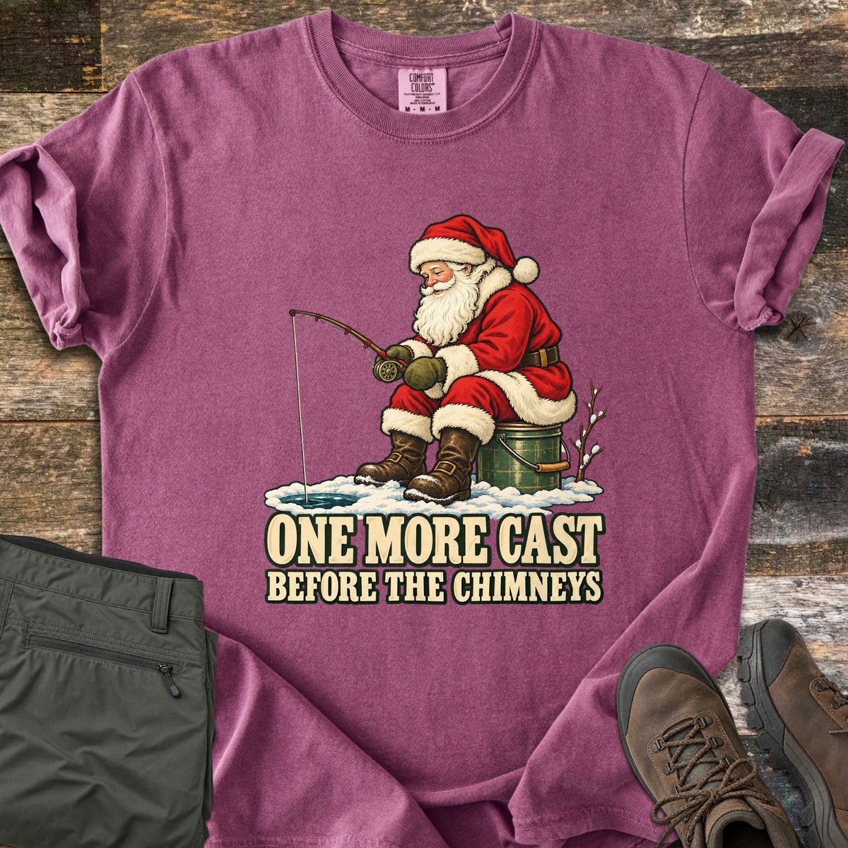 One More Cast Before the Chimneys Comfort Colors T-shirt in Berry, laid flat. Santa sits on a green bucket holding a fishing rod over a hole in the snowy ice, above the words One More Cast Before the Chimneys. Folded olive pants and brown hiking boots lie on weathered wood.