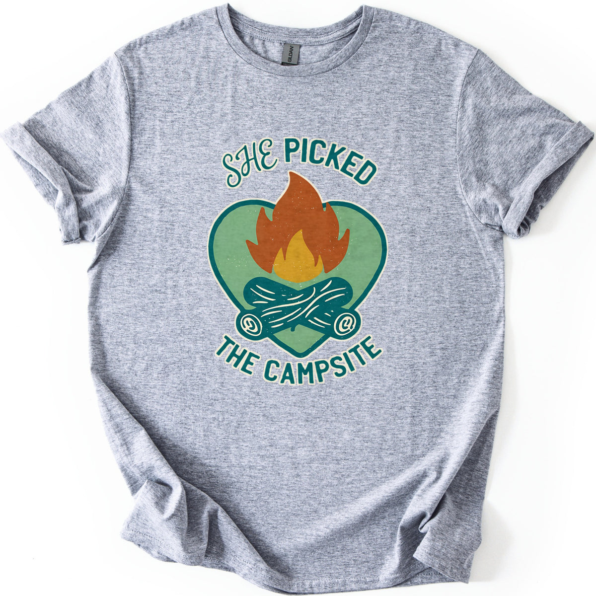 She Picked The Campsite Comfort Colors T-shirt in Sport Grey, laid flat. An orange and yellow campfire on crossed teal logs inside a sage green heart, with the words SHE PICKED above and THE CAMPSITE below. The shirt lies on a white surface with its sleeves cuffed.