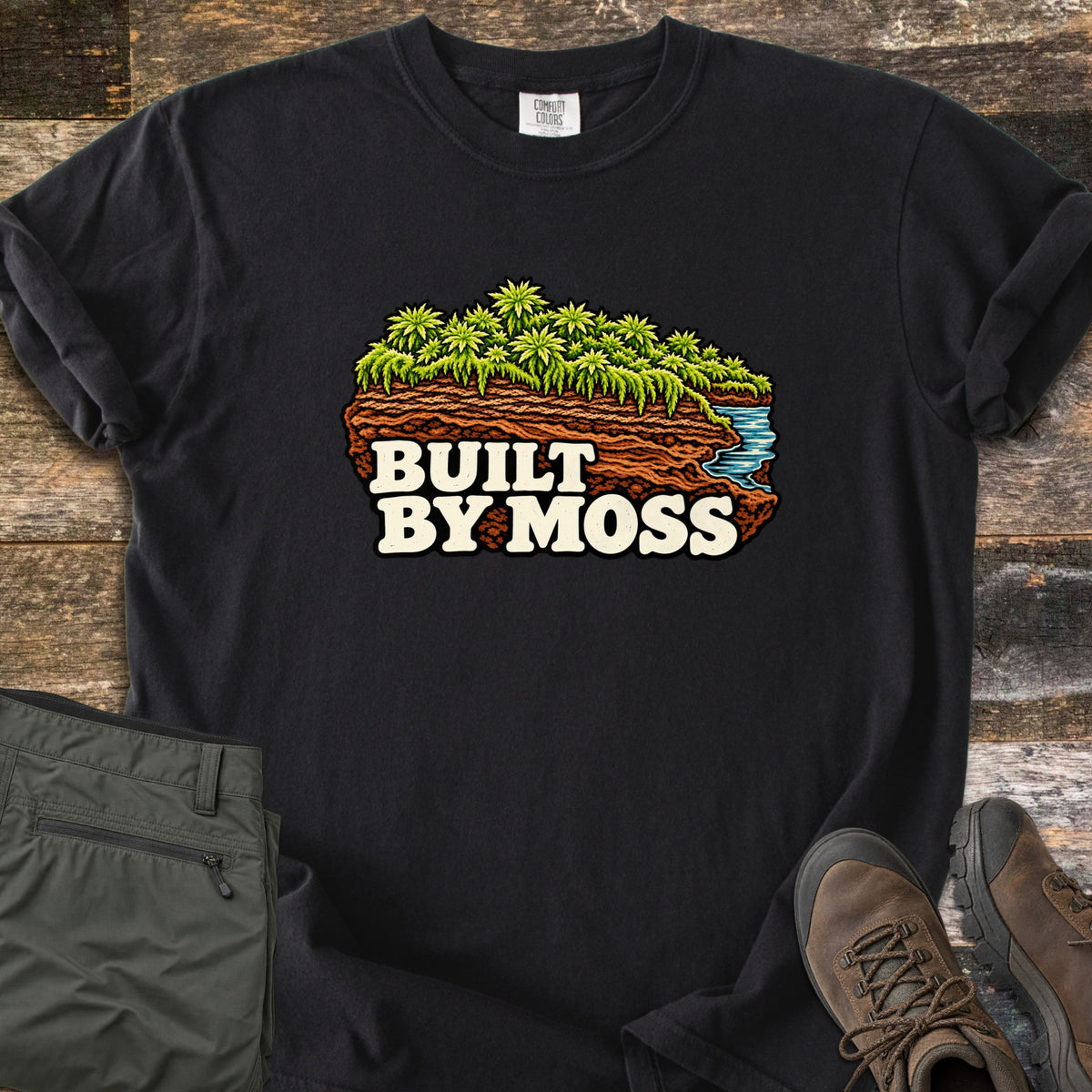Built by Moss Comfort Colors T-shirt in Black, laid flat. An illustrated chunk of brown soil topped with bright green moss and a small blue stream, with the words Built by Moss. Shown on weathered wood with folded olive hiking pants and brown hiking boots.