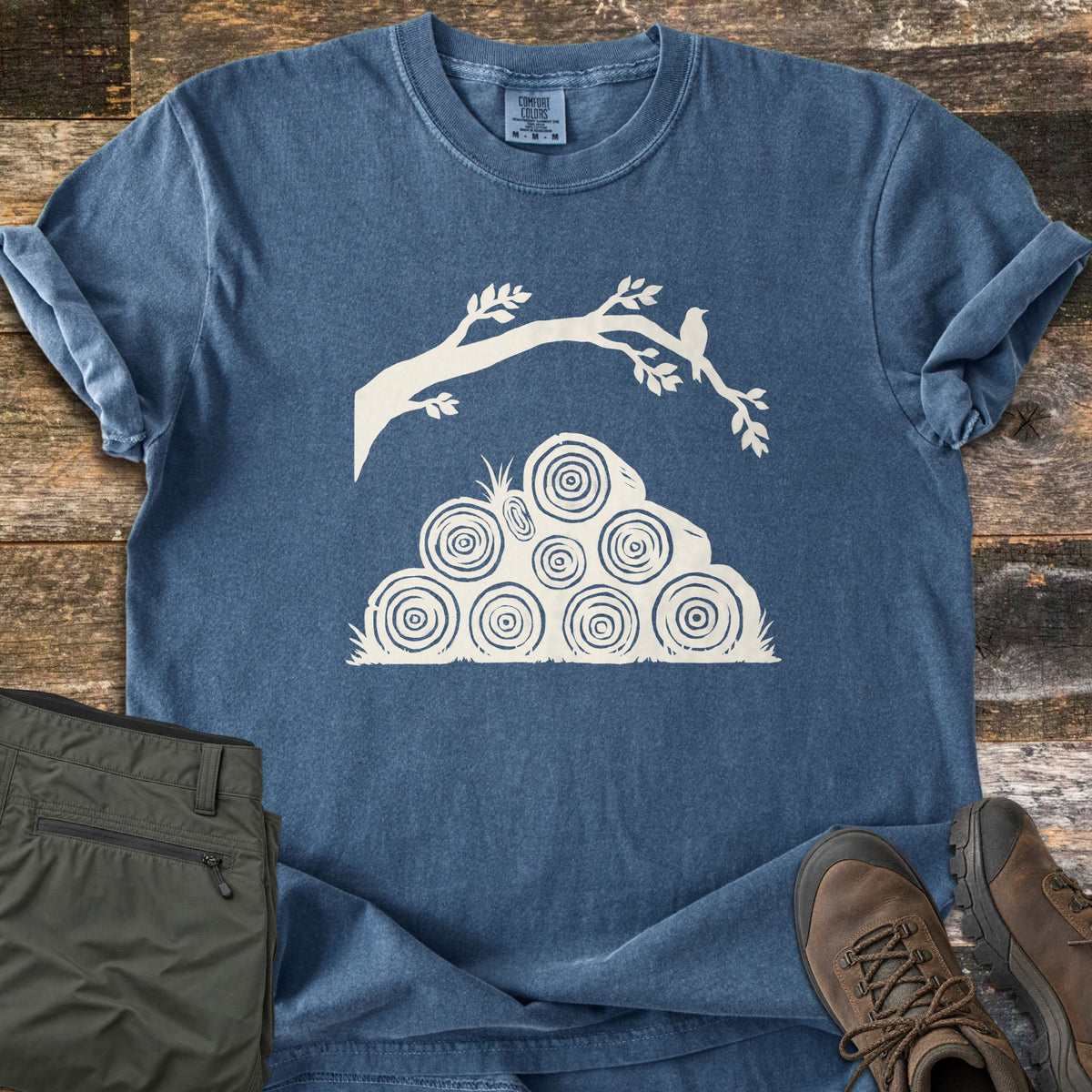 Stacked Firewood Comfort Colors T-shirt in Denim, laid flat. A cream print of stacked log rounds with tree rings under a leafy branch where a small bird perches. Sleeves are rolled on a weathered wood background beside olive hiking pants and brown hiking boots.