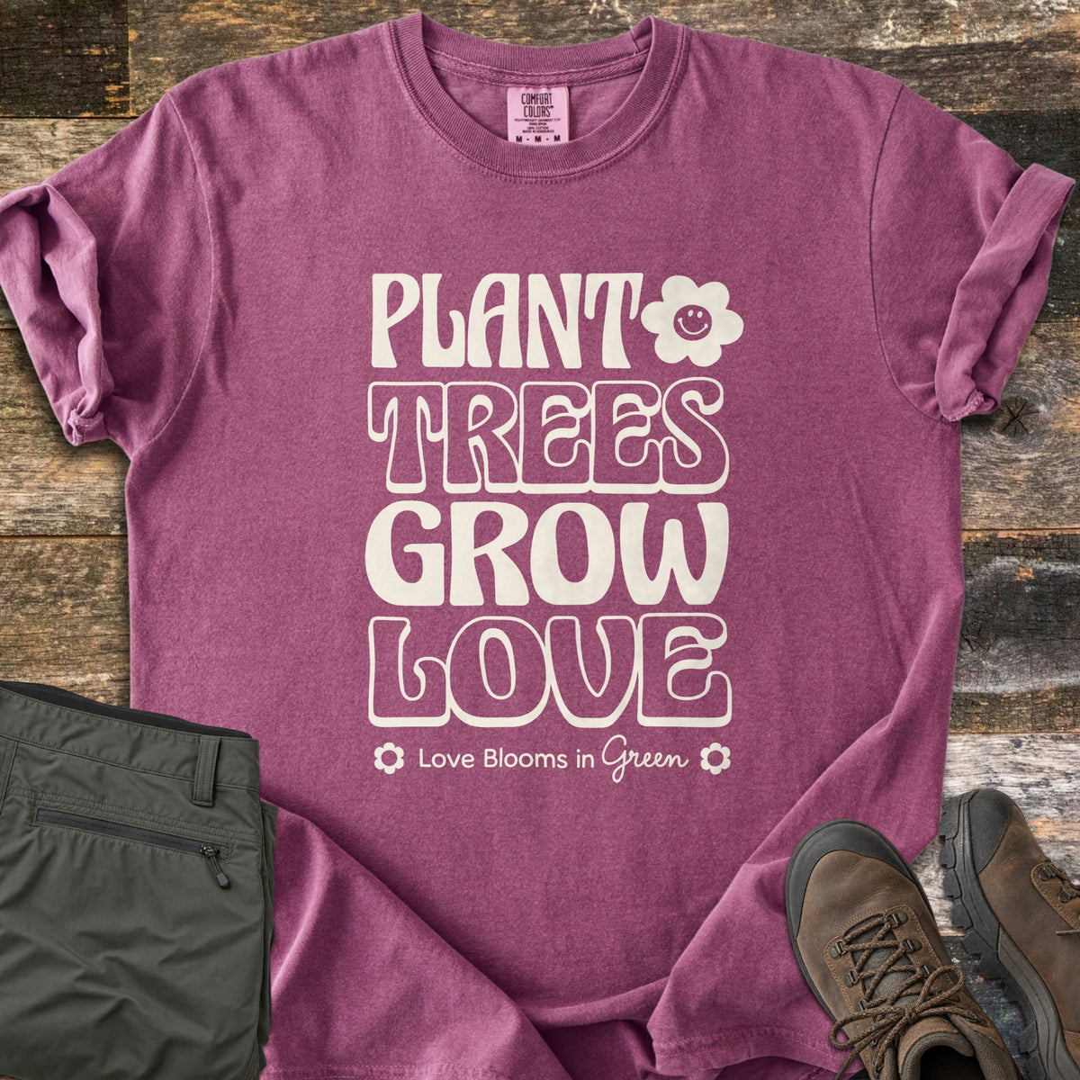 Plant Trees Grow Love Comfort Colors T-shirt in Berry, laid flat. Cream retro bubble letters read Plant Trees Grow Love beside a smiling daisy, above Love Blooms in Green. Sleeves cuffed on weathered wood with folded olive hiking pants and brown hiking boots.