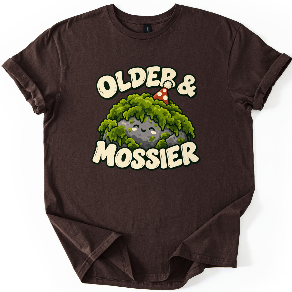 Older & Mossier Comfort Colors T-shirt in Dark Chocolate, laid flat. A smiling gray rock draped in green moss wears a red party hat with white dots, between the words Older & and Mossier in cream bubble letters. The shirt lies on a white background with its sleeves cuffed.