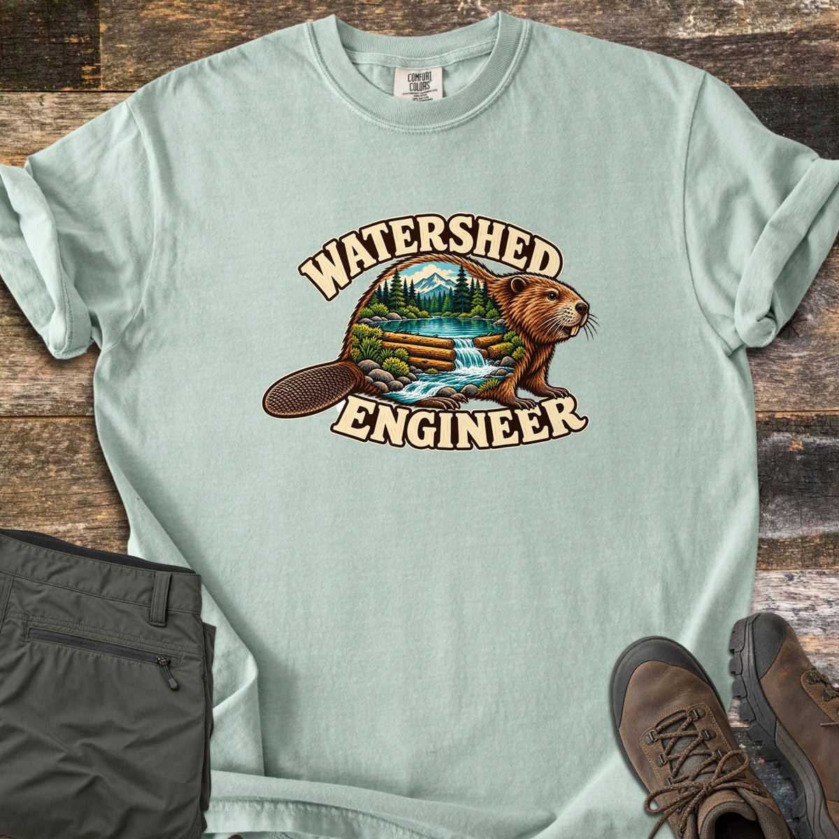 Watershed Engineer Comfort Colors T-shirt in Bay, laid flat. A beaver whose body frames a forest scene with mountains, a lake, and a waterfall flowing over a log dam, between the words Watershed Engineer. Olive pants and brown hiking boots lie on a wood surface.