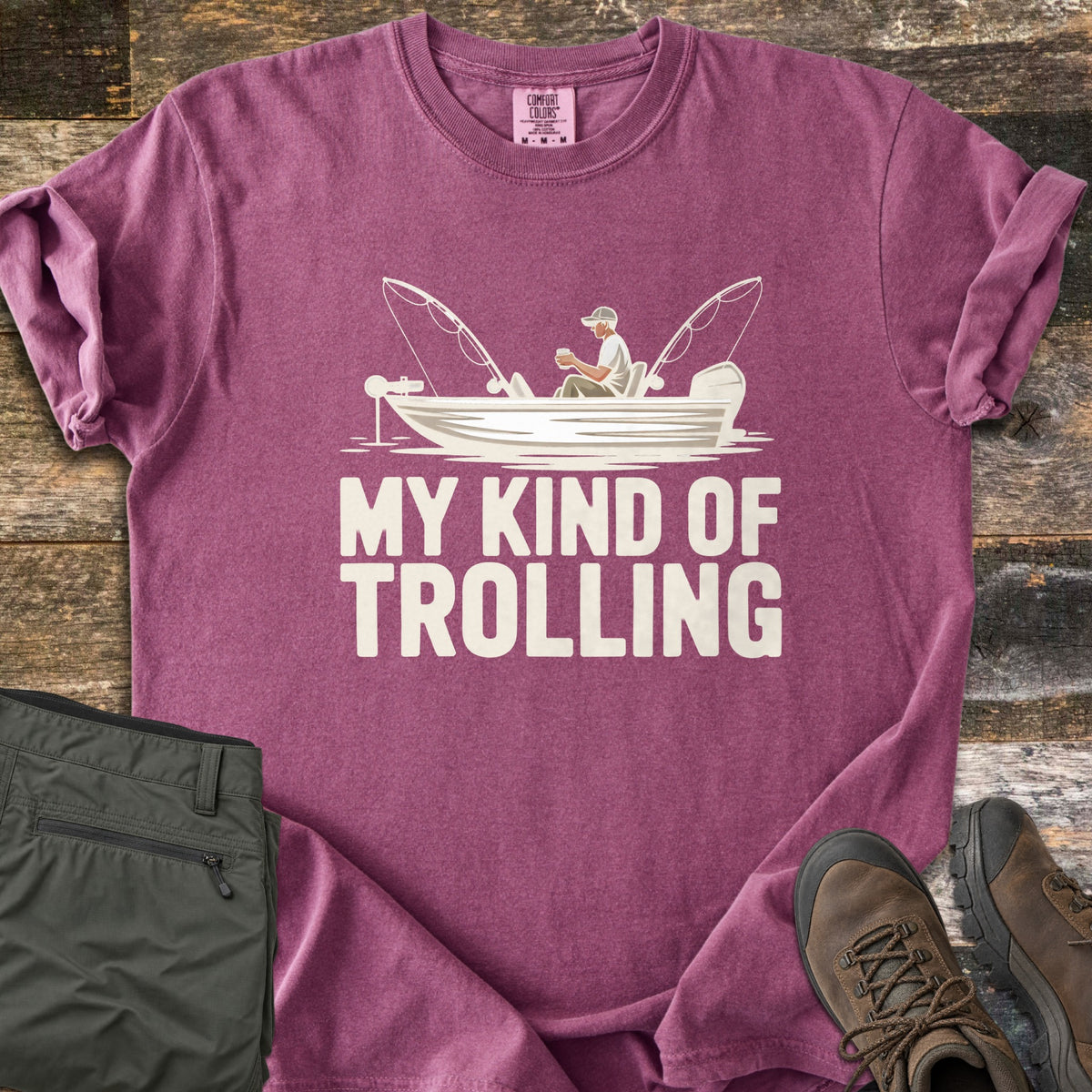 My Kind Of Trolling Comfort Colors T-shirt in Berry, laid flat. A man in a cap holds a cup in a small fishing boat with two bent rods and motors, above the words My Kind Of Trolling in cream. Olive cargo pants and brown hiking boots sit beside it on weathered wood.