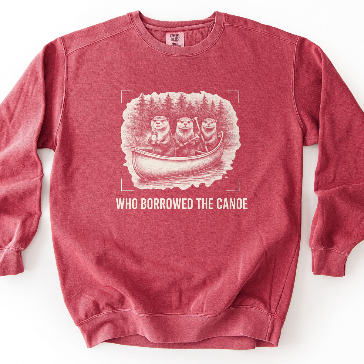 Who Borrowed The Canoe Comfort Colors crewneck sweatshirt in Crimson, laid flat. A cream sketch of three otters in a canoe on a lake with a pine forest behind, one holding a lantern, framed by corner brackets. Below it the words WHO BORROWED THE CANOE in cream. On a white surface.