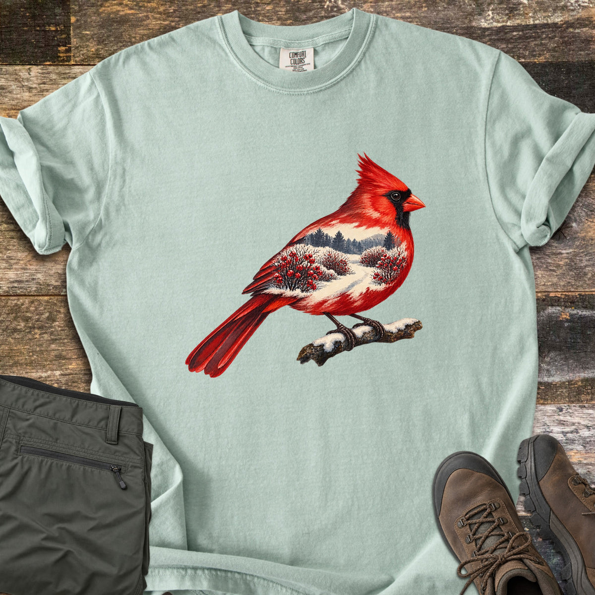 Winter Cardinal Comfort Colors T-shirt in Bay, laid flat. A red cardinal perched on a snowy branch, its body filled with a snowy landscape of pine trees and red berry bushes. Rolled sleeves on weathered wood, with olive pants and brown hiking boots beside it.