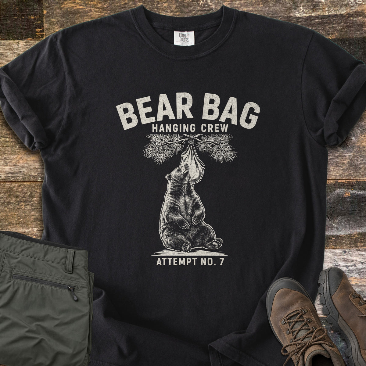 Bear Bag Hanging Crew Comfort Colors T-shirt in Black, laid flat. A sketched bear sits under a pine branch sniffing a hanging food bag, with the words Bear Bag Hanging Crew above and Attempt No. 7 below. Olive hiking pants and brown hiking boots lie beside it on weathered wood.